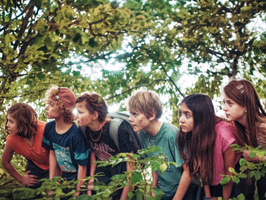 Auf dem Foto aus dem Film Die unlangweiligste Schule der Welt sind sechs Kinder im Wald zu sehen, die sich in Blättern verstecken und alle aufmerksam nach links blicken.