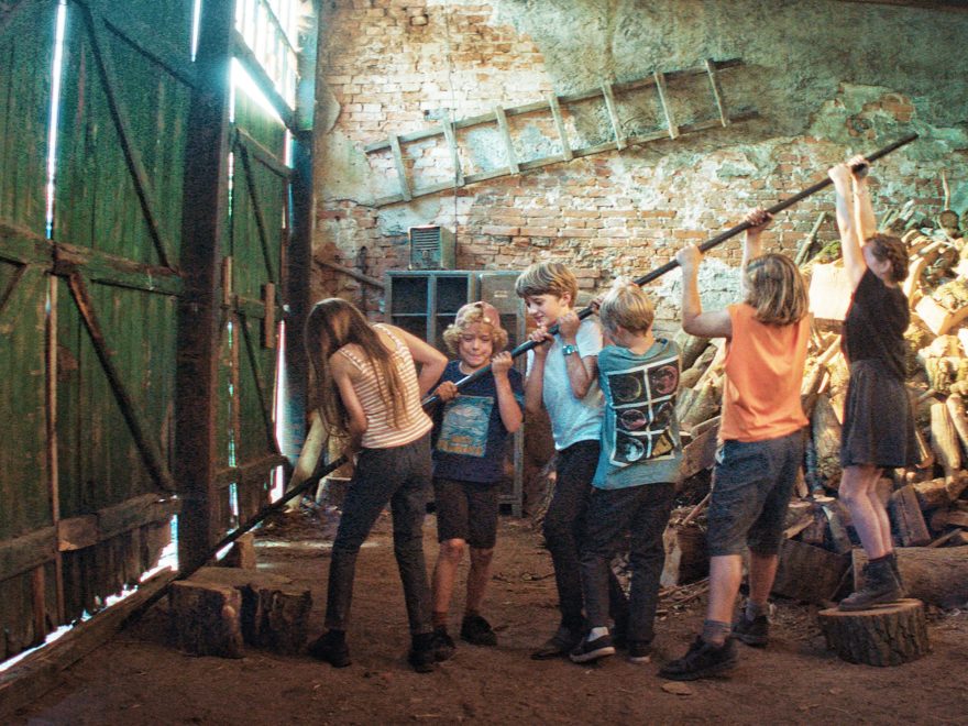 Maxe und seine fünf Klassenkameraden versuchen mithilfe einer langen Brechstange ein großes Scheunentor aus den Angeln zu heben. Im Hintergrund sind Regale und ein Haufen Feuerholz zu erkennen. An der Backsteinwand hängt eine Holzleiter.