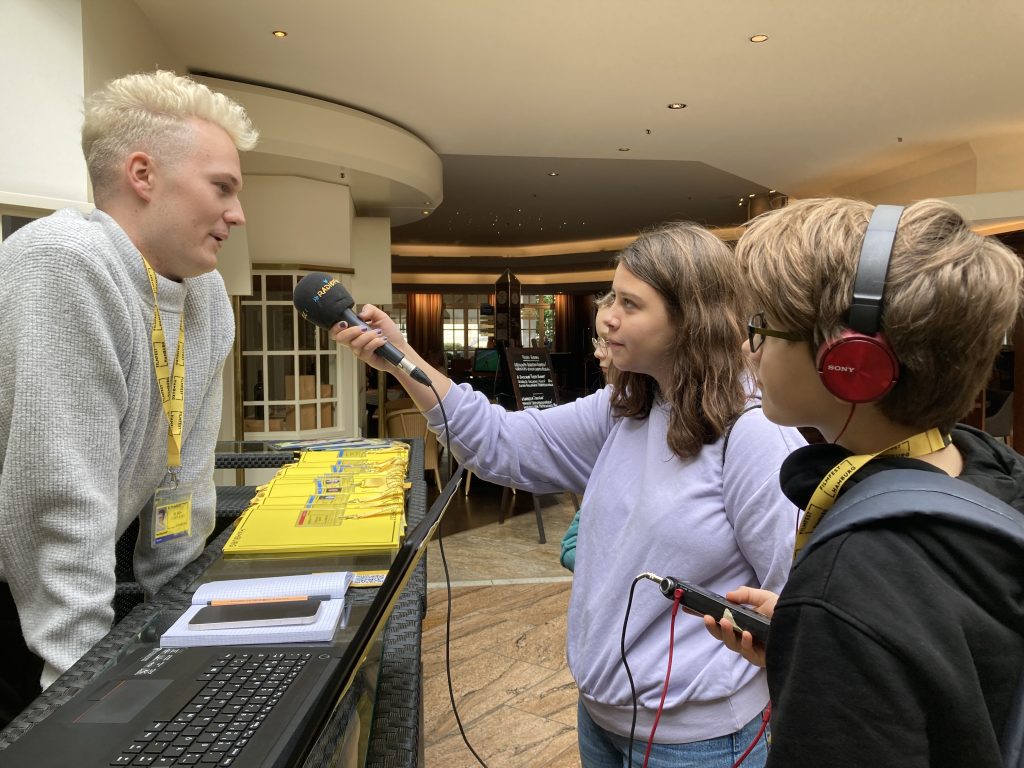 Zwei Jugendliche stehen nach links gedreht zu ihrem Interviewpartner. Die mittlere Person hält das Mikrofon. Links davon steht ein Mitarbeiter der Filmfests Hamburg. Sie befinden sich alle in der Lobby eines Hotels.