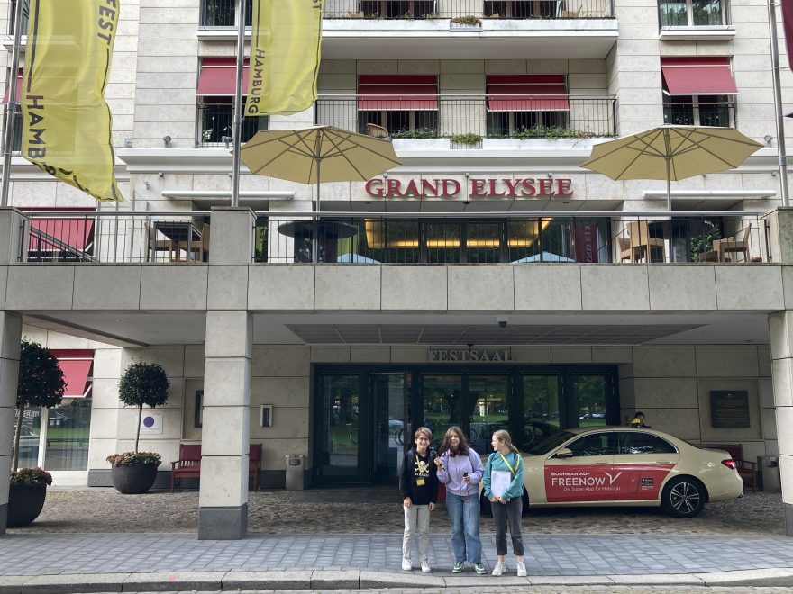 Drei Jugendliche stehen vor einem Hotel. Vor dem unteren Eingang steht ein Taxi. Oberhalb stehen aufgespannte Sonnenschirme. Die roten Rollläden und die gelben Fahnen des Filmfests Hamburg sind außerdem sichtbar.
