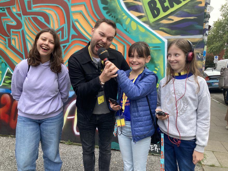 Auf dem Bild stehen ein Erwachsener und drei Kinder vor einer bunten Graffiti-Wand. Sie blicken fast alle in die Kamera. Der Erwachsene tut so, als würde er in ein Mikrofon beißen.