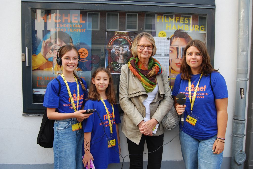 Auf dem Foto sind die MICHEL MOVIE KIDS Svea, Yara und Lenja zu sehen vor dem Studio Kino zu sehen - in der Mitte steht die Buchautorin Kirsten Boie. Im Hintergrund sind Kinoplakate zu sehen.