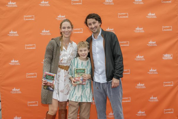 Auf dem Foto sind die Autorin Sabrina Kirschner, der Regisseur Ekrem Ergün und der junge Schauspieler Lucas Herzog zu sehen. Im Hintergrund sieht man die Logos vom MICHEL Kinder und Jugend Filmfest.